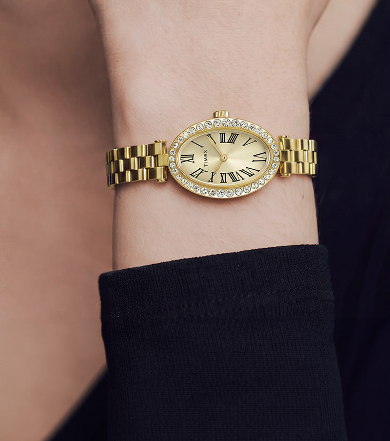 Reloj Timex Cavatina® Crystals TW2Y90100 en tono dorado, con esfera ovalada color champán y bisel adornado con cristales, lucido en una muñeca en relieve junto a una manga negra.