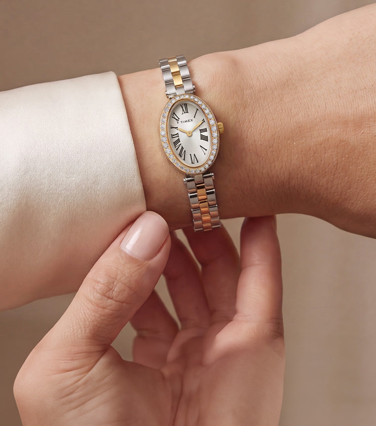 TW2Y90200: Reloj Timex Cavatina® Crystals en dos tonos, con bisel ovalado adornado con cristales y esfera con números romanos, lucido junto al puño de una blusa de color marfil.