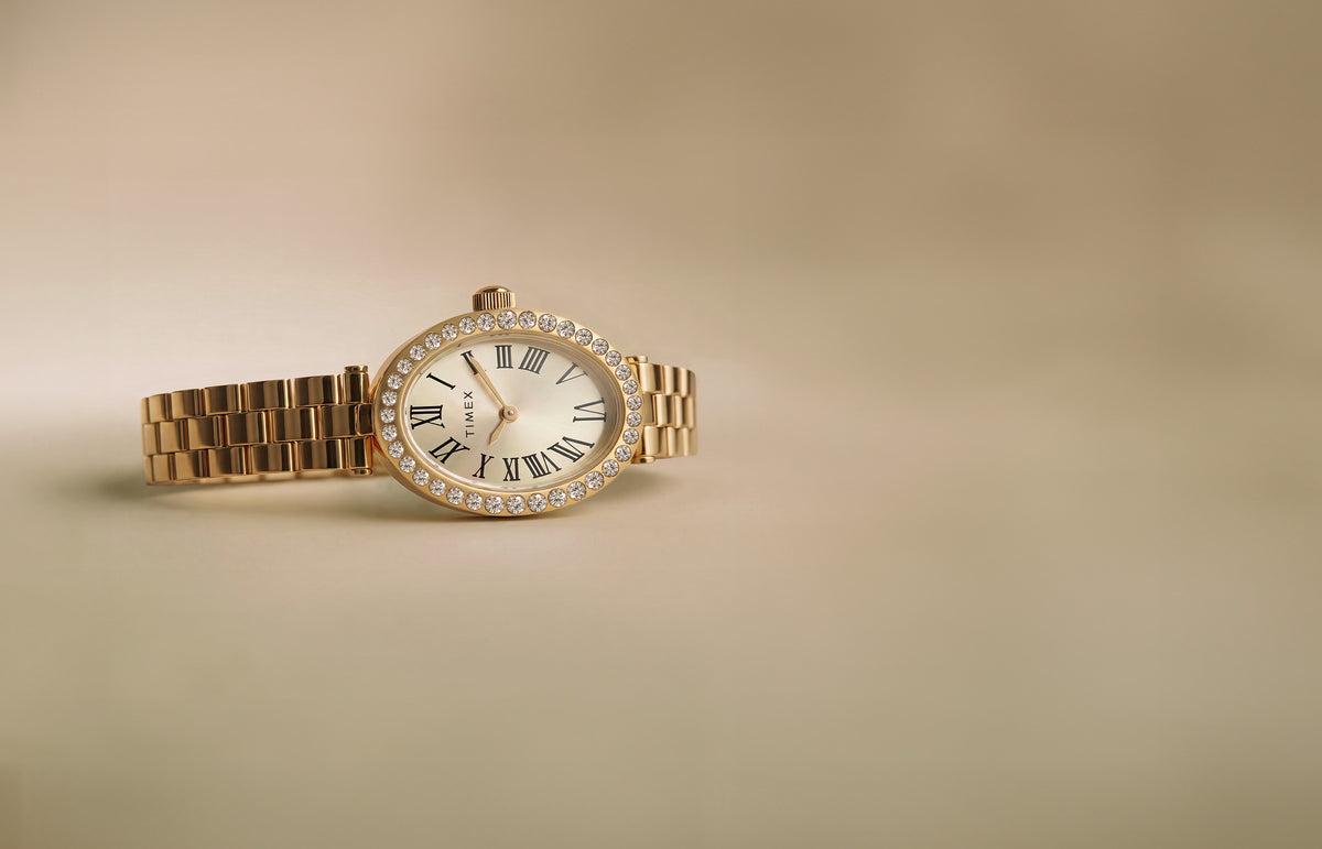 Reloj Timex Cavatina® Crystals TW2Y90100 con brazalete en tono dorado, esfera ovalada color champán y bisel adornado con cristales sobre fondo beige.