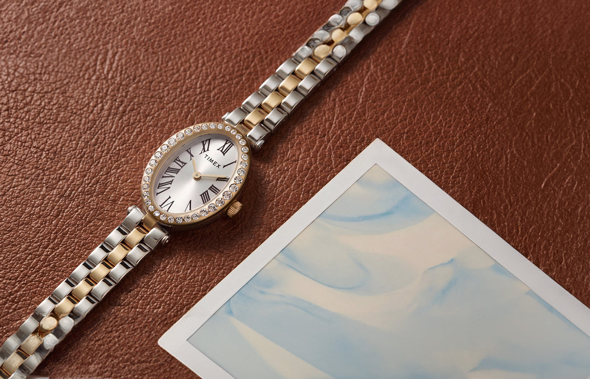 Reloj Timex Cavatina® Crystals TW2Y90200 con brazalete bicolor, esfera ovalada en tono plateado y bisel dorado con incrustaciones de cristales, junto a una fotografía de cuero marrón.