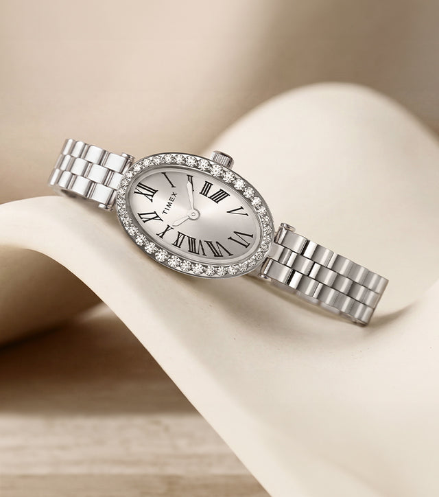 Reloj Timex Cavatina® Crystals TW2Y90000 con esfera ovalada con números romanos, bisel con cristales y brazalete en tono plateado, presentado sobre un expositor curvo de color marfil.