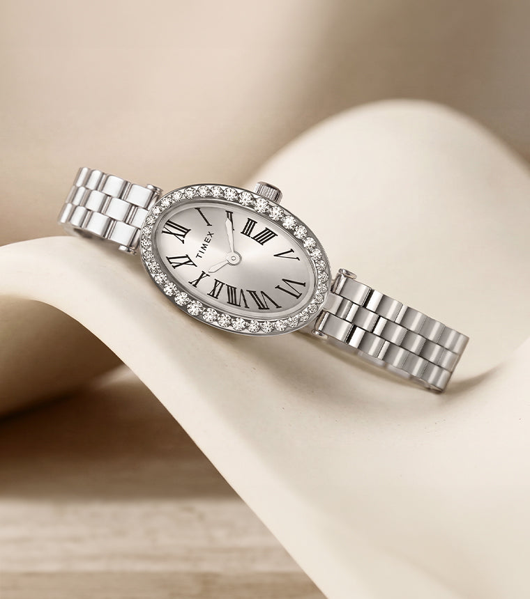 Reloj Timex Cavatina® Crystals TW2Y90000 con esfera ovalada con números romanos, bisel con cristales y brazalete en tono plateado, presentado sobre un expositor curvo de color marfil.