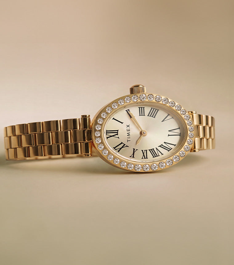 Reloj Timex Cavatina® Crystals TW2Y90100 con brazalete en tono dorado, esfera ovalada color champán y bisel adornado con cristales sobre fondo beige.