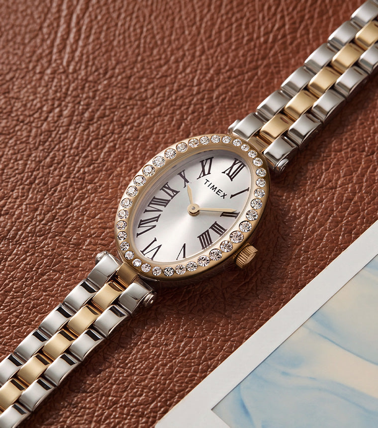 Reloj Timex Cavatina® Crystals TW2Y90200 con brazalete bicolor, esfera ovalada en tono plateado y bisel dorado con incrustaciones de cristales, junto a una fotografía de cuero marrón.