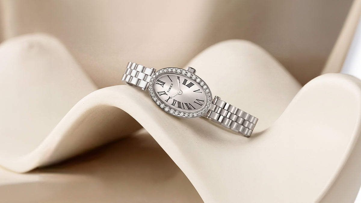 Reloj Timex Cavatina® Crystals TW2Y90000 en tono plateado, presentado en un expositor de color marfil con forma de ola, con una esfera ovalada y un bisel bordeado de cristales.