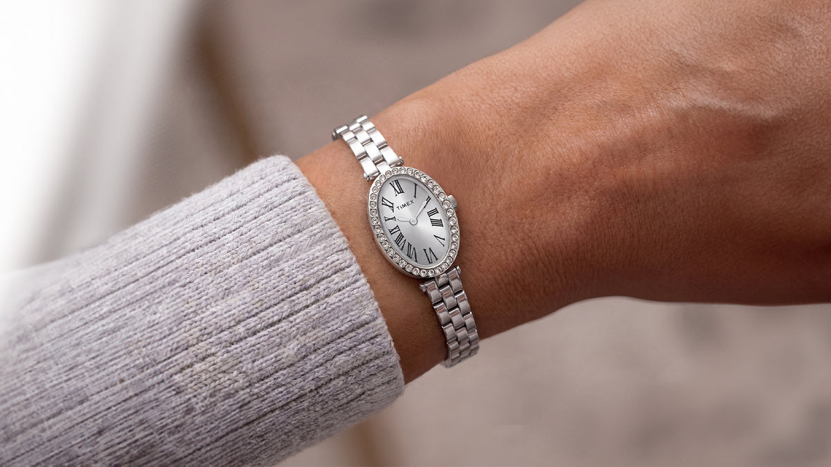 Reloj Timex Cavatina® Crystals modelo TW2Y90000 en una muñeca, en el que se aprecia su caja ovalada bordeada de cristales y su fina pulsera plateada junto a un jersey gris.