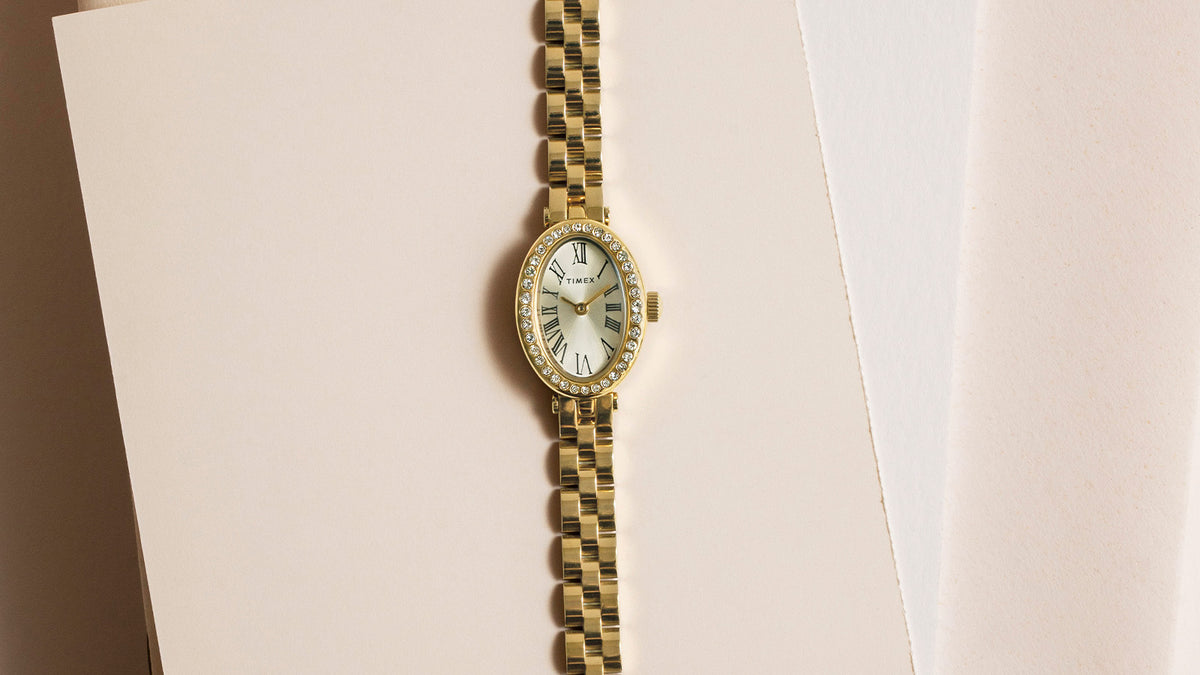 Reloj Timex Cavatina® Crystals TW2Y90100 en tono dorado, con esfera ovalada con números romanos y bisel con cristales, colocado sobre papel color crema.