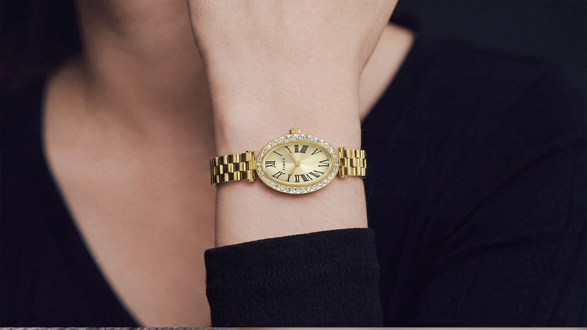 Reloj Timex Cavatina® Crystals (TW2Y90100) lucido en la muñeca, con una pulsera de eslabones en tono dorado y una caja ovalada con cristales incrustados, a juego con un conjunto negro.