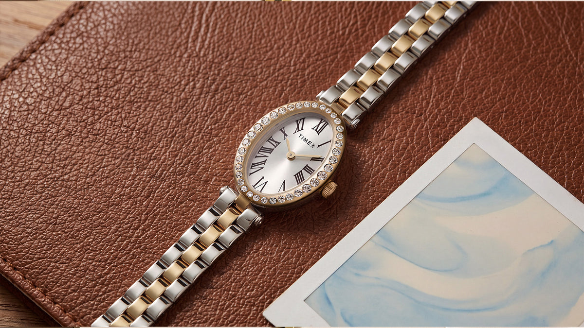 Reloj Timex Cavatina® Crystals (TW2Y90200) con brazalete de eslabones en tonos plateados y dorados, apoyado en diagonal sobre una correa de piel marrón junto a una fotografía.