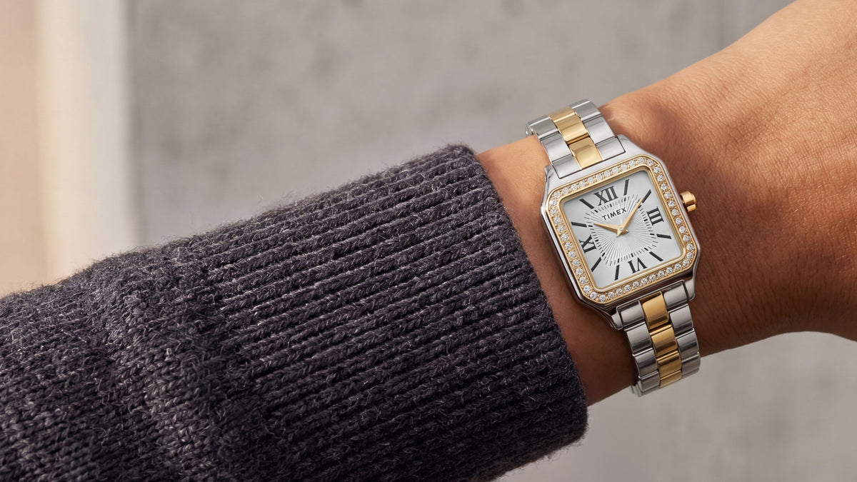 Reloj Timex Addison TW2Y90400 con brazalete en tonos plateados y dorados, esfera blanca con números romanos y bisel con incrustaciones de cristal, combinado con un jersey de punto color carbón.