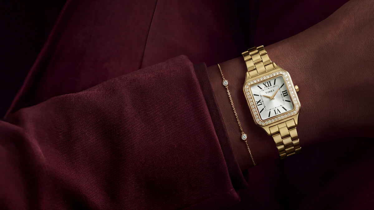 TW2Y90500 Gold-tone Timex Addison watch with a crystal-lined bezel and white Roman numeral dial, styled with a delicate bracelet and burgundy outfit.