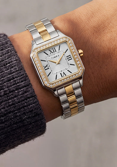 Reloj Timex Addison TW2Y90400 con brazalete en tonos plateados y dorados, esfera blanca con números romanos y bisel con incrustaciones de cristal, combinado con un jersey de punto color carbón.
