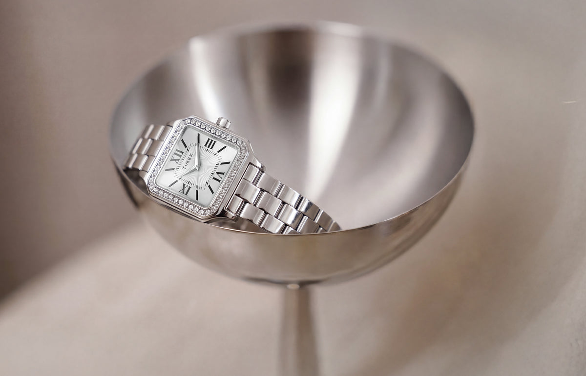 TW2Y90300 Timex Addison watch with a silver-tone bracelet, white Roman numeral dial, and crystal-lined bezel, resting in a metal dish.