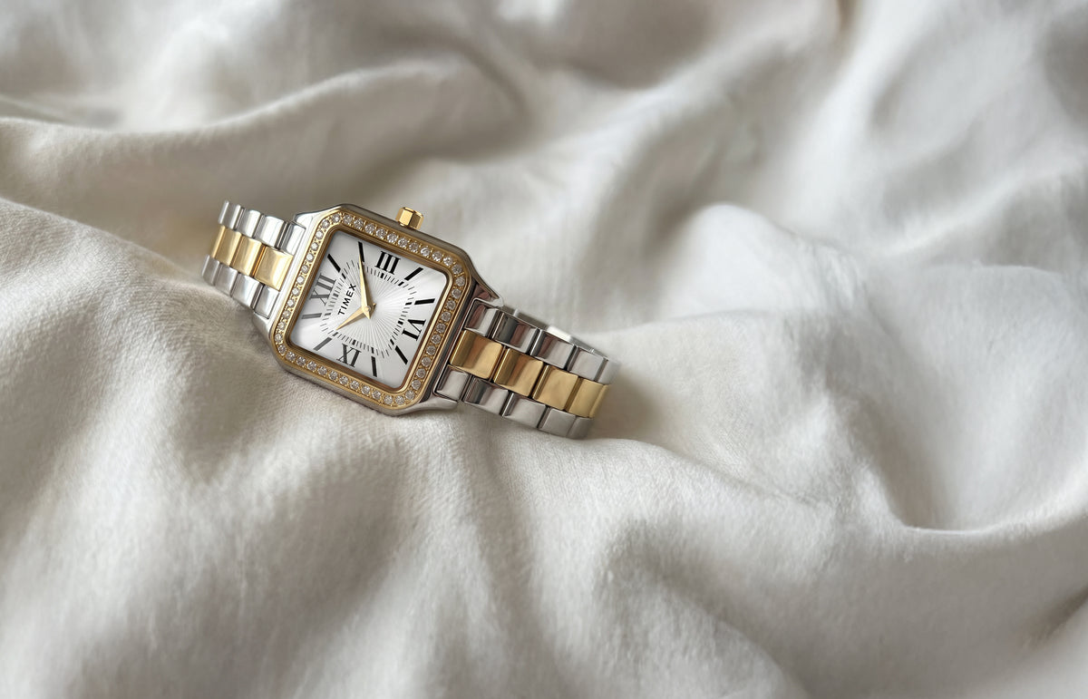 Reloj Timex Addison TW2Y90400 con brazalete en tonos plateados y dorados, esfera blanca con números romanos y bisel con incrustaciones de cristal, sobre una tela de color marfil.