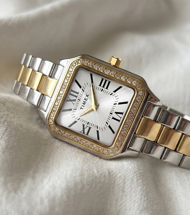 Reloj Timex Addison TW2Y90400 con brazalete en tonos plateados y dorados, esfera blanca con números romanos y bisel con incrustaciones de cristal, sobre una tela de color marfil.