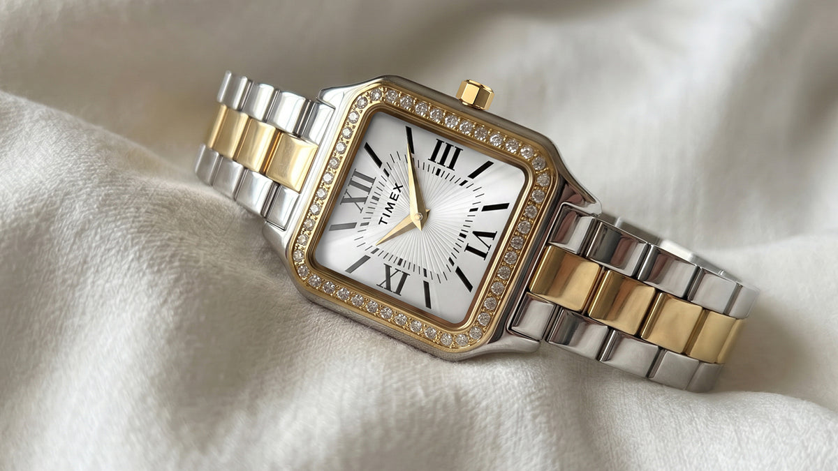 TW2Y90400 Primer plano del reloj Timex Addison sobre un tejido de color marfil, en el que se aprecia su bisel con incrustaciones de cristales y su brazalete de eslabones pulidos en tonos plateados y dorados.
