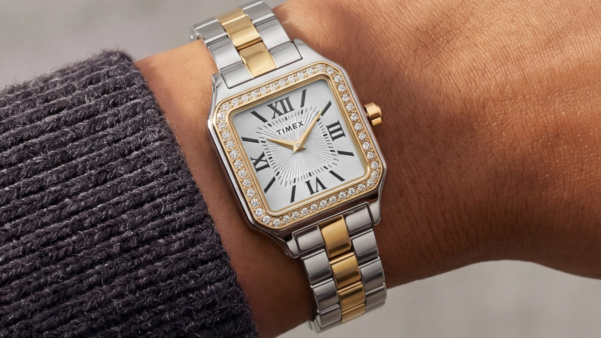 TW2Y90400 Primer plano del reloj Timex Addison bicolor en una muñeca, con agujas doradas y un bisel con incrustaciones de cristal, junto a una manga de punto color carbón.
