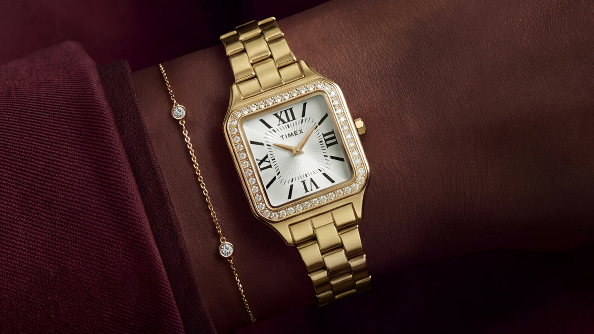 TW2Y90500 Gold-tone Timex Addison watch worn on a wrist beside a delicate gold-tone bracelet and burgundy sleeve.