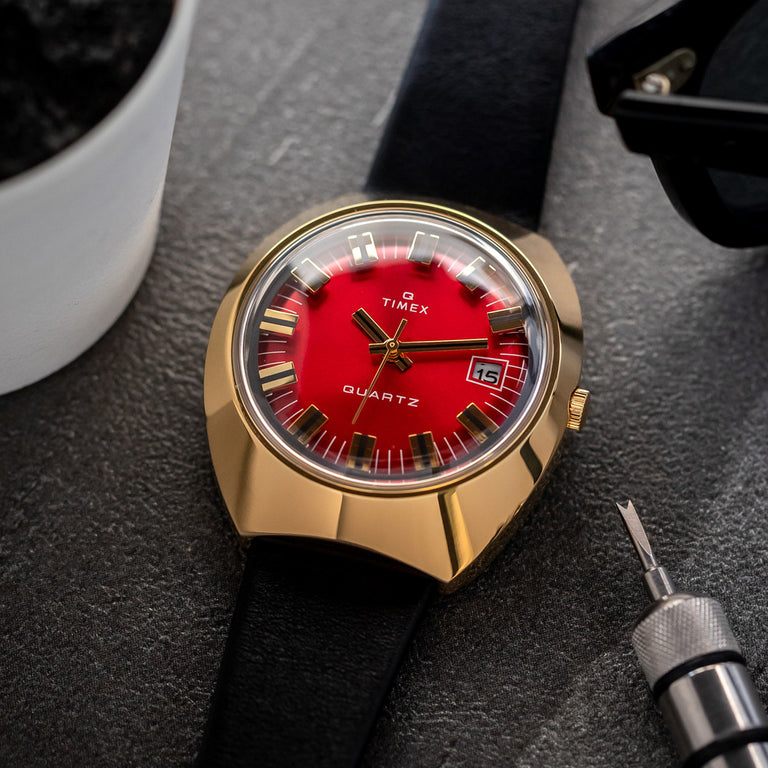 Timex Q Reissue watch with a bold red dial, gold-tone case, black leather strap, and date window, displayed on a textured surface with watchmaking tools and accessories.