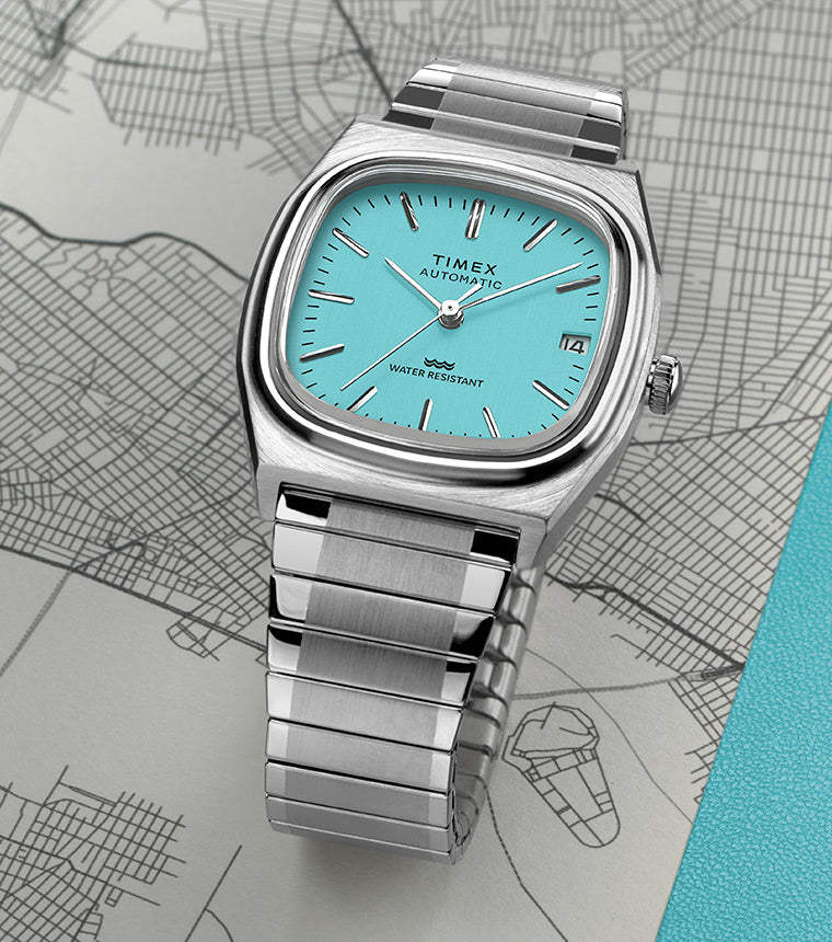 Timex® Automatic 1983 E Line watch with a turquoise dial and stainless-steel bracelet resting on a city map.