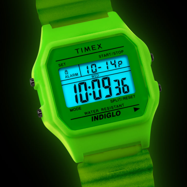 Close-up of a bright green Timex T80 digital watch with a blue INDIGLO® backlight against a dark background.
