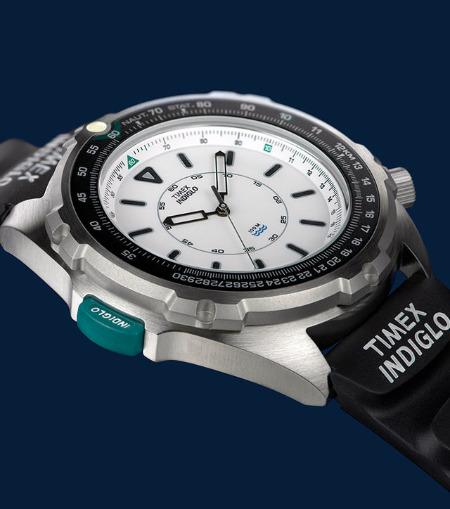 A detailed close-up of the Timex 1995 Intrepid Reissue watch, showcasing its brushed stainless steel case, black rotating bezel, and crisp white dial with black indices and hands. The black rubber strap features the 'TIMEX INDIGLO' branding, and a teal INDIGLO button is prominently visible. The image has a dark blue background, emphasizing the watch's design and materials.