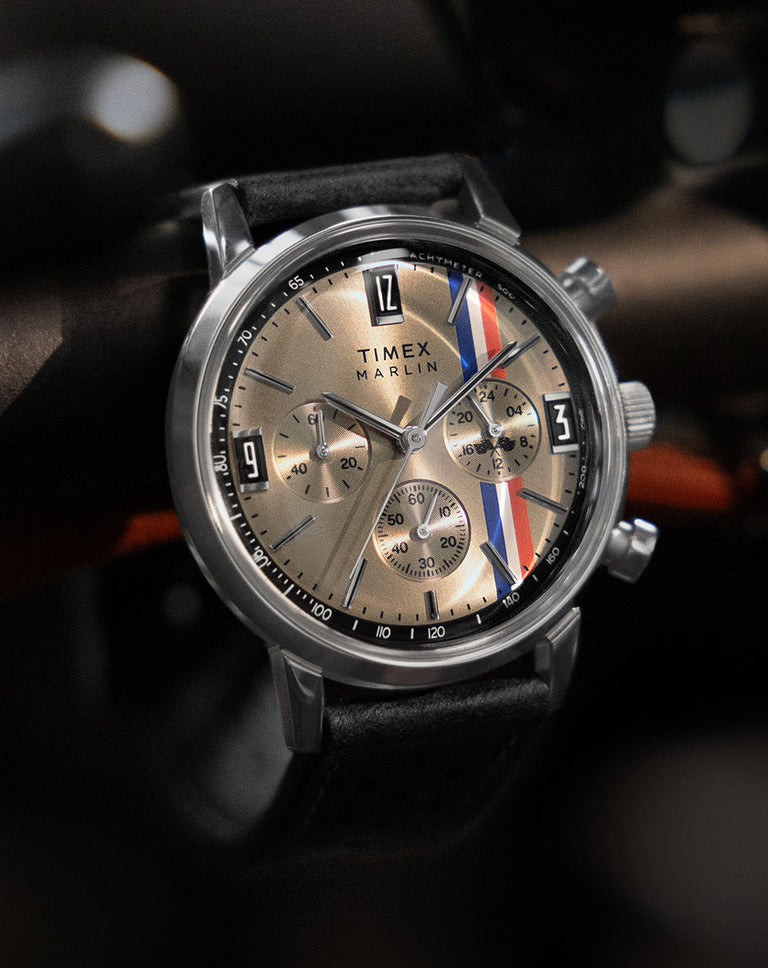 A close-up shot of the Timex Marlin® Chronograph watch, featuring a champagne-colored dial with a vintage-inspired tachymeter scale and bold racing stripes in red, white, and blue. The stainless steel case and chronograph subdials enhance its classic motorsport aesthetic, while the black leather strap adds a refined touch. The watch is set against a dark, blurred background, emphasizing its sleek and sophisticated design.