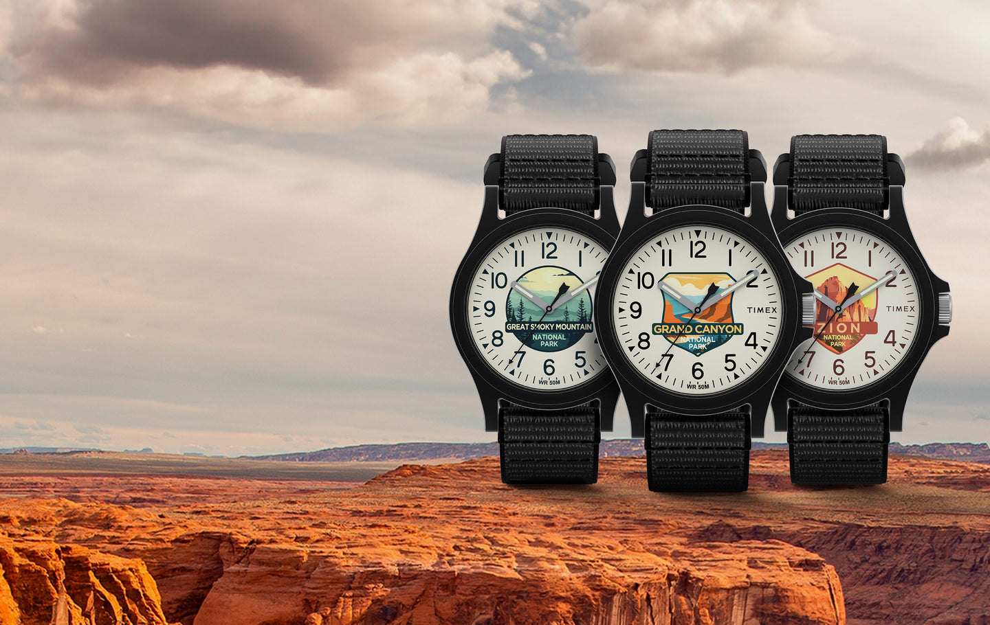 Timex Watch with black straps and colorful dials featuring illustrations of Great Smoky Mountain National Park, Grand Canyon National Park, and Zion National Park, displayed against a scenic background of a rocky canyon landscape under a cloudy sky.