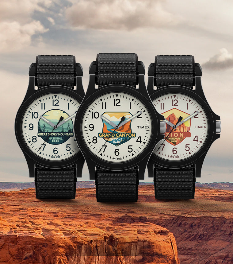 Timex Watch with black straps and colorful dials featuring illustrations of Great Smoky Mountain National Park, Grand Canyon National Park, and Zion National Park, displayed against a scenic background of a rocky canyon landscape under a cloudy sky.