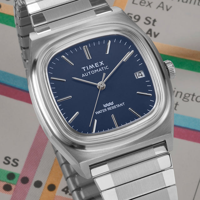 A close-up of a Timex E-Line Automatic wristwatch with a deep blue dial, minimalist silver hour markers, and a date display. The watch features a sleek, brushed stainless steel case and bracelet, evoking a refined and modern aesthetic. It is set against a background of a New York City subway map, symbolizing urban style and functionality.