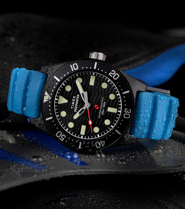 TW2Y02500 Timex Deepwater 200M dive watch with a blue silicone strap resting on wet diving gear. The black dial features bold luminous markers, wavy texture, and a red seconds hand. Water droplets emphasize its 200-meter water resistance and rugged design, ideal for underwater use.