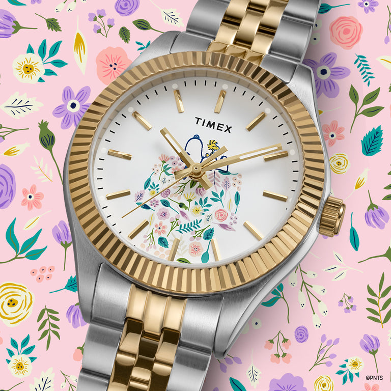 Timex watch with a two-tone gold and silver bracelet, white dial featuring Snoopy surrounded by colorful floral designs, displayed on a matching pastel floral background.