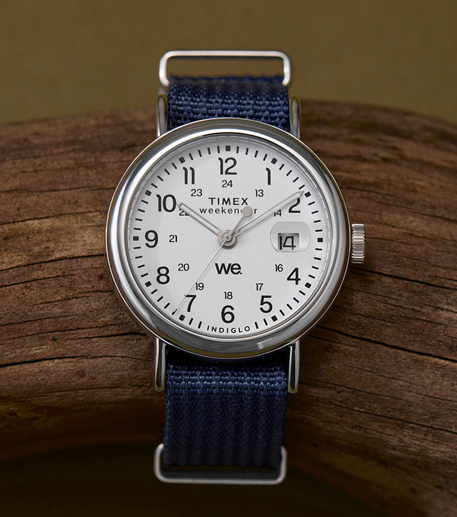 A Timex Weekender 37mm watch with a white dial and navy blue fabric strap, resting on a piece of driftwood. The dial features black Arabic numerals, 24-hour military time, and a date magnifier at 3 o’clock.