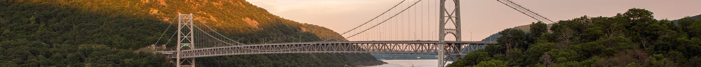 A scenic view of the Bear Mountain Bridge stretching over the Hudson River, framed by lush green mountains during sunset, with soft golden light hitting the landscape.