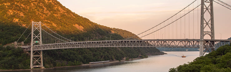 A scenic view of the Bear Mountain Bridge stretching over the Hudson River, framed by lush green mountains during sunset, with soft golden light hitting the landscape.