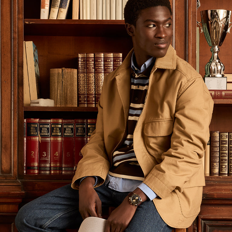 A stylish young man sits in front of a classic wooden bookshelf filled with leather-bound books and a trophy. He wears a tan jacket over a striped sweater and blue jeans, showcasing a bold wristwatch on his left wrist.