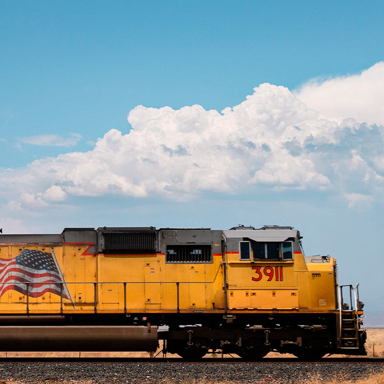 A bright yellow freight train engine with the number 3911 painted in red on the side travels along a track in a flat, arid landscape. The side of the engine features a large, waving American flag decal. Above, a vivid blue sky is filled with fluffy white clouds, creating a striking contrast with the yellow of the train.