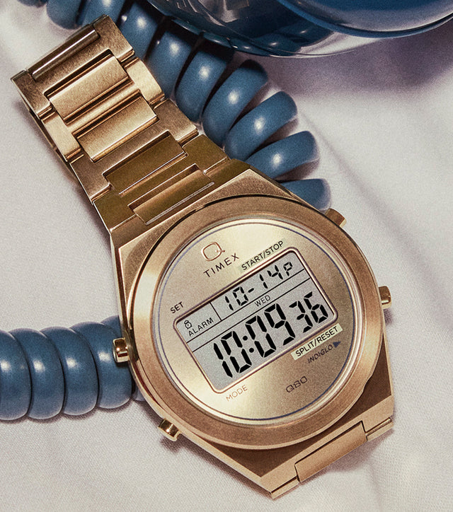A gold-tone Q Timex® Q80 Continental digital watch resting on a white bedsheet, wrapped in the coiled cord of a vintage blue rotary telephone.