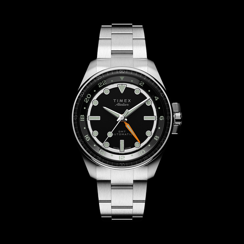 Product display of the Timex Atelier GMT Automatic in stainless steel with a black dial and orange GMT hand, styled for marketing layout.