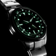 The Timex Atelier GMT Automatic illuminated in low light, highlighting the glowing green dial and orange GMT hand for nighttime legibility.