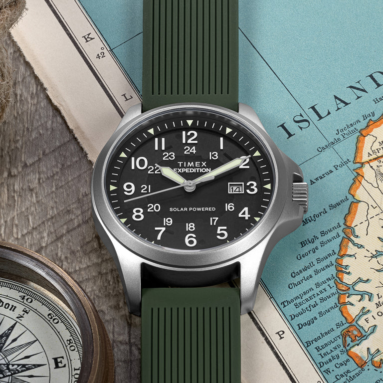 "Timex Expedition Solar Powered watch with a black dial, luminous hands, silver-tone case, and green textured strap. The dial features both 12-hour and 24-hour markers along with a date window at 3 o’clock. The watch is placed on a vintage map with a compass, highlighting its rugged outdoor adventure style.