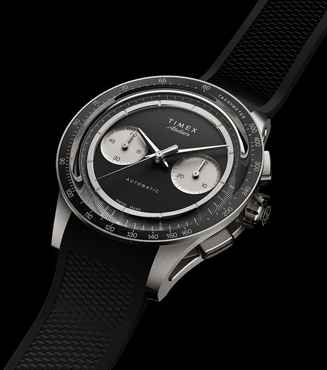 Timex Atelier Chronograph Automatic M1a Ti titanium watch with black dial, silver-tone sub-dials, tachymeter bezel, and textured black rubber strap shown at an angled perspective.