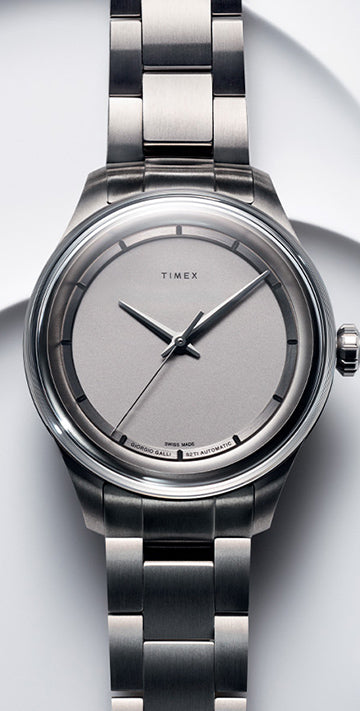 The image shows a sleek, minimalist wristwatch by Timex. The watch has a simple, round dial in a muted silver color, with no numeric markers—only minimalist indices at the hour positions. The design is elegant, with a polished finish and clean lines, reflecting a modern and sophisticated aesthetic.