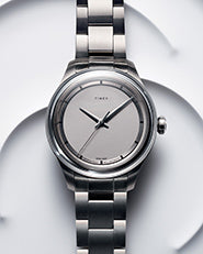 The image shows a sleek, minimalist wristwatch by Timex. The watch has a simple, round dial in a muted silver color, with no numeric markers—only minimalist indices at the hour positions. The design is elegant, with a polished finish and clean lines, reflecting a modern and sophisticated aesthetic.