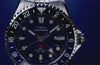 Timex Deepwater Meridian GMT Automatic watch with a blue dial and two-tone 24-hour bezel rotates against a blue background, highlighting its stainless-steel case, red GMT hand, and crown.