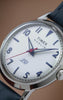 Close-up video of the Timex Marlin® America 250 watch with a white dial, blue leather strap, and red seconds hand, rotating through angled and front-facing views against a warm brown background to highlight its polished stainless-steel case and vintage-inspired design.