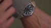 Person wearing and adjusting the MK1® Automatic watch with a black dial and tan fabric strap.