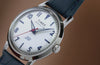 Close-up video of the Timex Marlin® America 250 watch with a white dial, blue leather strap, and red seconds hand, rotating through angled and front-facing views against a warm brown background to highlight its polished stainless-steel case and vintage-inspired design.