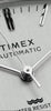Video showcasing the Timex® Automatic 1983 E Line Reissue watch (model TW2W70800) rotating on a map-themed background with yellow accents. The footage highlights the brushed stainless-steel case and integrated bracelet, silver sunburst dial with minimalist hour markers, and date window at 3 o’clock.