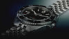 Promotional video showcasing the Timex Deepwater Arctic watch in close-up detail. The stainless steel dive watch with a black dial and rotating bezel appears against dark blue lighting, with rotating angles highlighting the bracelet, face, and textured bezel. Intercut underwater scenes show scuba divers swimming above coral reefs and schools of fish, reinforcing the watch’s ocean-inspired, professional diving aesthetic.